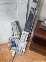 Photo showing towel bars, hooks, closet pole and assorted hardware in packaging set on floor near furniture.