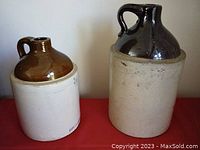 Both antique two-tone stoneware whiskey/moonshine jugs shown side by side on a red surface. Larger jug on right has brown glazed neck and handle, beige stoneware body. Smaller jug on left has similar design with lighter brown glaze.