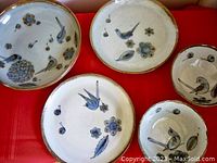 Top-down view of all five stoneware ceramics laid out on a red surface showing hand-painted bird and flower designs on cream base.