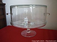 Crystal cake stand with cover shown assembled on red surface, clear glass with two side handles on cover.