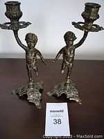 Pair of metal cherub candle holders standing side by side on a surface, viewed from the front to show full height and detail.
