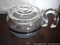 Profile view showing clear glass body of teapot with black band and curved handle.