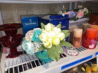 Shelf view of Royal Doulton figurines in blue boxes, artificial flowers in blue-green vase, decorative candles, and part of ruby red candy dish.