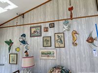 Wide view of sun porch wall showing multiple pieces of beach and outdoor themed wall art including framed needlepoint pictures and various wooden carvings of birds, fish, and aquatic animals.