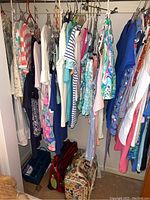 Wide view of hanging summer clothing in closet with various colorful tops and dresses on hangers, including white, blue, and floral prints. Bags visible on floor.