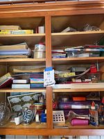 Wide view of wooden shelf with multiple compartments filled with office supplies including paper, ribbon, tape, stapler boxes, glue bottle, and assorted stationery.