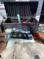 Black outdoor storage container open to show items inside including cushions, statues, a red leaf blower, doormats, and cords.