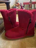 Side profile showing the red suede material, plush red fur lining visible at collar, black side zipper with branded puller, and flat brown sole.