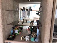 Wide view of three glass shelves containing various animal figurines including cats, dogs, and others. Several items have fluffy or textured fur look.