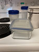 Photo showing ceramic canisters, plastic zip-lock containers with blue lids, and clear glass container with plastic lid on stove.