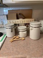 Three white ceramic kitchen canisters of varying sizes with metal clasp lids and three wooden measuring spoons in front, original cardboard box in the background.