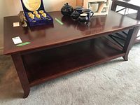 Rectangular wood veneer coffee table with lower shelf, polished dark finish, positioned on carpeted floor with decorative items on top. Visible minor scratches and chips.
