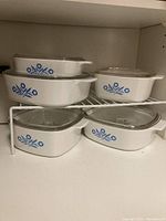 Five white Corning Ware casseroles with Blue Cornflower design arranged on a shelf, each with clear Pyrex glass lids.
