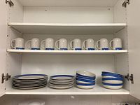 Open white cabinet shelf showing 8 mugs with blue and gray band design, 7 dinner plates stacked, 8 lunch plates stacked, and 8 bowls stacked