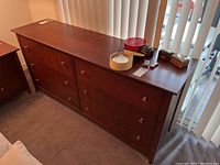 Six drawer wooden dresser seen from an angle showing six drawers with silver metal knobs, reddish brown finish, and some non-included items on top.