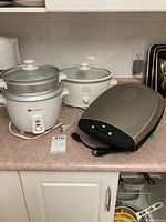 Three kitchen appliances on countertop: White-Westinghouse rice cooker with steaming tray on top, Rival slow cooker, and George Foreman grill.