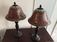 Pair of decorative metal and glass table lamps with purple and gold bell-shaped shades and dark metal bases with intricate carvings