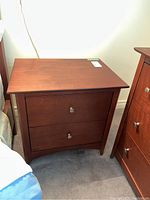 Full front view of wooden nightstand showing two drawers with metal pull knobs, sitting on carpeted floor beside a bed and dresser.