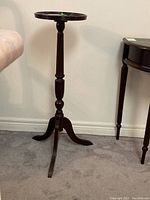 Front view of dark wood pedestal plant stand with slender turned column and circular top, tripod curved legs on carpeted floor.