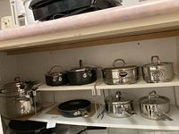 Two shelves of various pots and pans including stainless steel pieces and non-stick frying pans, all with lids except frying pans.