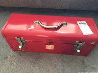 Closed red metal toolbox with handle and latch locks on carpeted floor.
