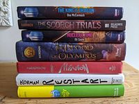 Stack of seven hardcover books showing titles and authors on spine, including Doctor Who, The Scorch Trials, Nuclear Time, and The Blood of Olympus.