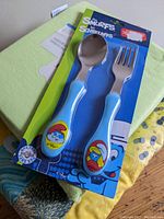 Close-up of Smurfs baby cutlery set in retail packaging, includes spoon and fork with blue handles and Smurf character images
