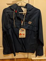 Full front view of navy blue vintage windbreaker jacket on hanger showing hood, half-zip with white zipper, attached original hang tag, and Rally Express logo patch on chest.