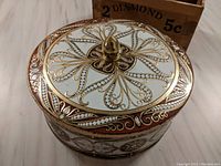 Oval vintage tin with intricate gold and white decorative design and finial knob on top