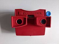 Red 3D View-Master toy showing front facing twin lenses, embossed logo on top and blue lever on side.