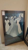 Full view of framed poster leaning against a wall, showing abstract dancing figures in white and blue tones with text visible at bottom.