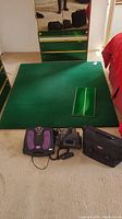 Photo showing two green golf mats on the floor in front of mirror, one smaller mat resting on larger mat. Purple and black electric foot massager with control panel, black massager device, carrying case placed on carpet next to mats.