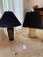 Pair of two black and gold table lamps with black shades on beige carpet near window with sheer white curtains.