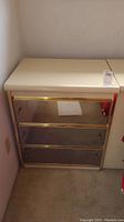 Nightstand viewed on an angle showing glossy cream top and three mirrored drawers with gold trims.