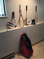 Five rackets including tennis, badminton, and racquetball types arranged against a wall, with a red and black carrying bag on the floor.