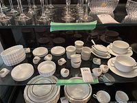Photo showing multiple white ceramic serving dishes such as ramekins, bowls, plates, and small cups. Positioned on glass shelves under crystal glassware.