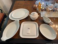 Photo showing a set of white ceramic serving ware on wooden table, including two oval platters, square CorningWare dish, oval bowl, butter dish with lid, cream pitcher.