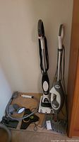 Two Shark steam mops standing upright along a wall. Black/white and gray/white models with cords wrapped around handles. Several mop pads and accessories scattered on the floor next to them.