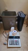 Photo shows gray metal trash bin, black electric paper shredder and Sharp printing calculator with paper rolls spaced on carpeted floor.