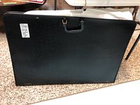 Closed black leather art case with handle and label 'Artista', placed on carpeted floor.