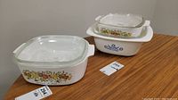 Three Corning Ware casserole dishes on wooden table, one Blue Cornflower design and two with vegetable pattern, all with lids.