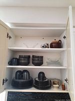 Full view of cabinet shelves showing stacked stoneware plates, mugs, glass plates and bowls, stoneware teapot, and small dishes.