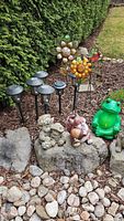 Photo showing six black solar garden lights, metal flower stakes, green frog garden figurine, chipmunk figurine, and stone gnome figurine arranged on garden stones and mulch.