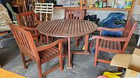 Full view of outdoor wooden dining table and four matching chairs arranged around it indoors on a concrete floor.