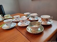 Wide angle photo showing all seven teacups with saucers on wooden table, showcasing variety of floral patterns and shapes.