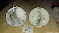 Two ceramic cups and saucers displayed side by side on a yellow surface. One set has blue and gold floral details, the other has green and multicolored floral blossoms.
