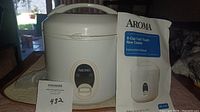 Photo of white Aroma 8-cup rice cooker with instruction manual alongside, placed on a dark surface.
