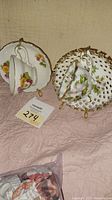 Photo showing two decorative china cup and saucer sets featuring floral designs with gold trim, one with pink and yellow flowers and one with green leaves and lattice edge saucer