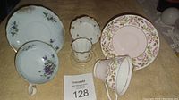 Five pieces of porcelain cups and saucers with floral patterns, placed on a beige surface with auction number 128.