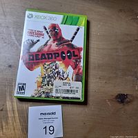 Full view of Deadpool Xbox 360 game case on wooden surface with auction tag showing lot number 19